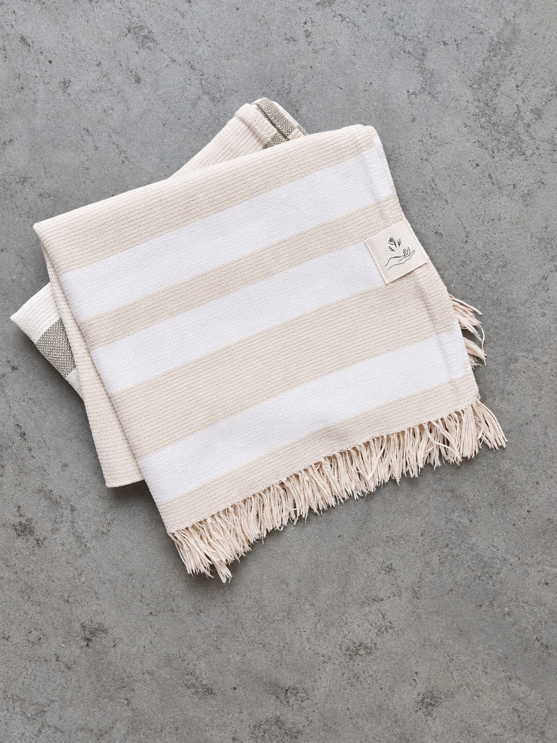A folded striped cotton Turkish towel with cream and white horizontal stripes, finished with fringed edges and a small woven brand label, displayed on a textured gray surface.