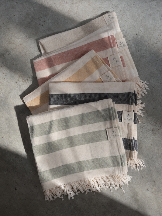 A stack of striped cotton towels with fringed edges, displayed on a textured concrete surface under natural sunlight. The towels feature stripes in sage green, dusty pink, mustard yellow, and dark charcoal gray on an off-white base.