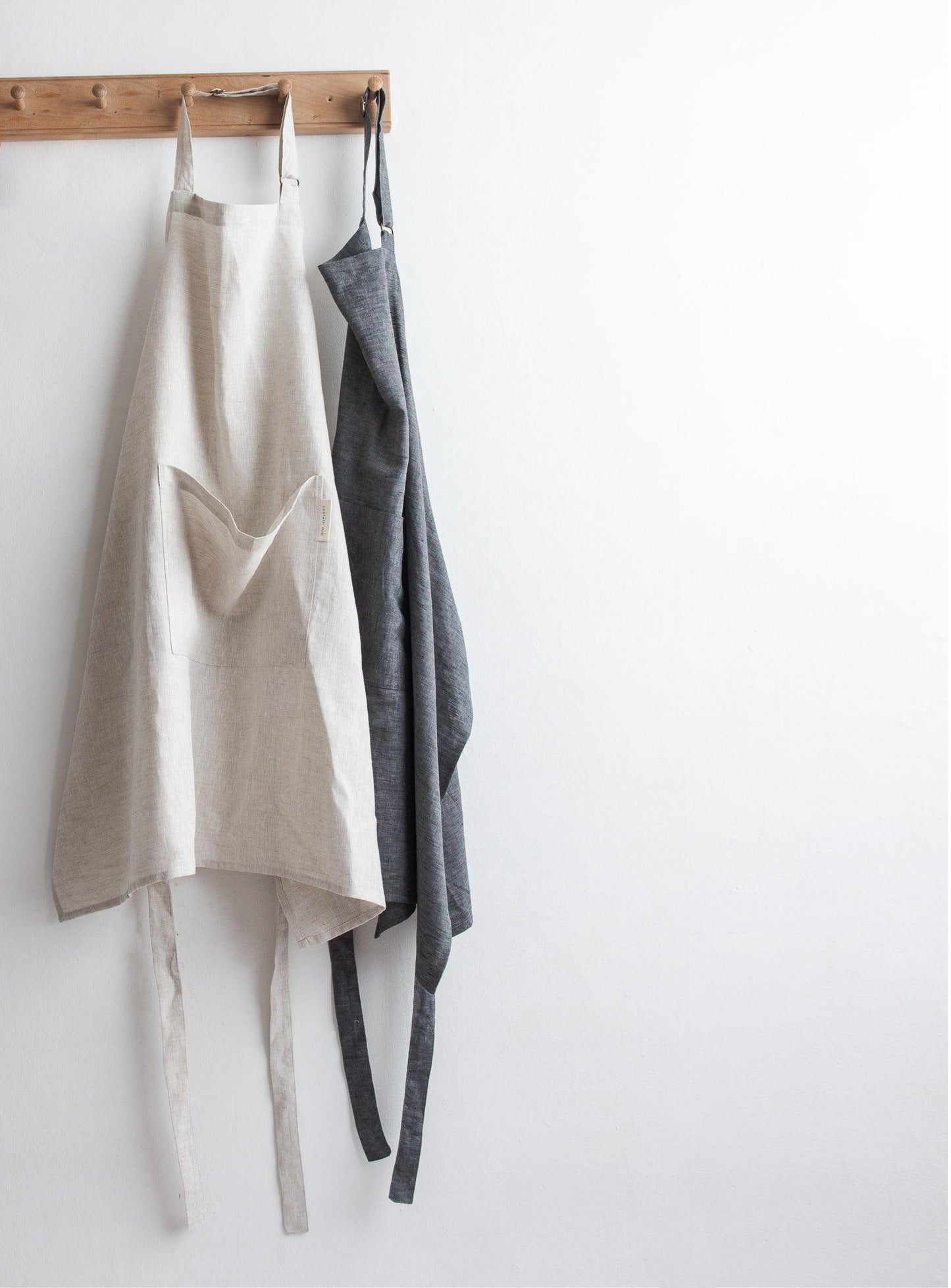 one beige and one grey apron hung on a wooden peg rail