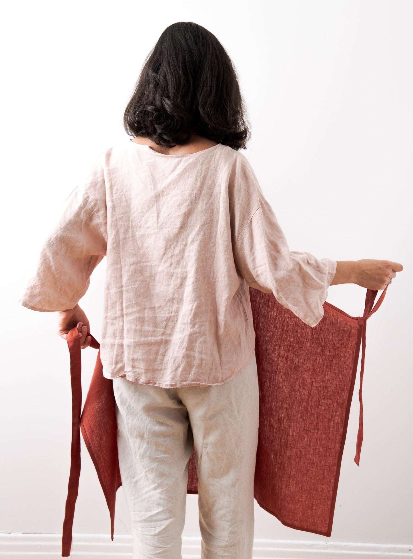 woman in pink shirt and linen pants seen from behind, putting on a red apron