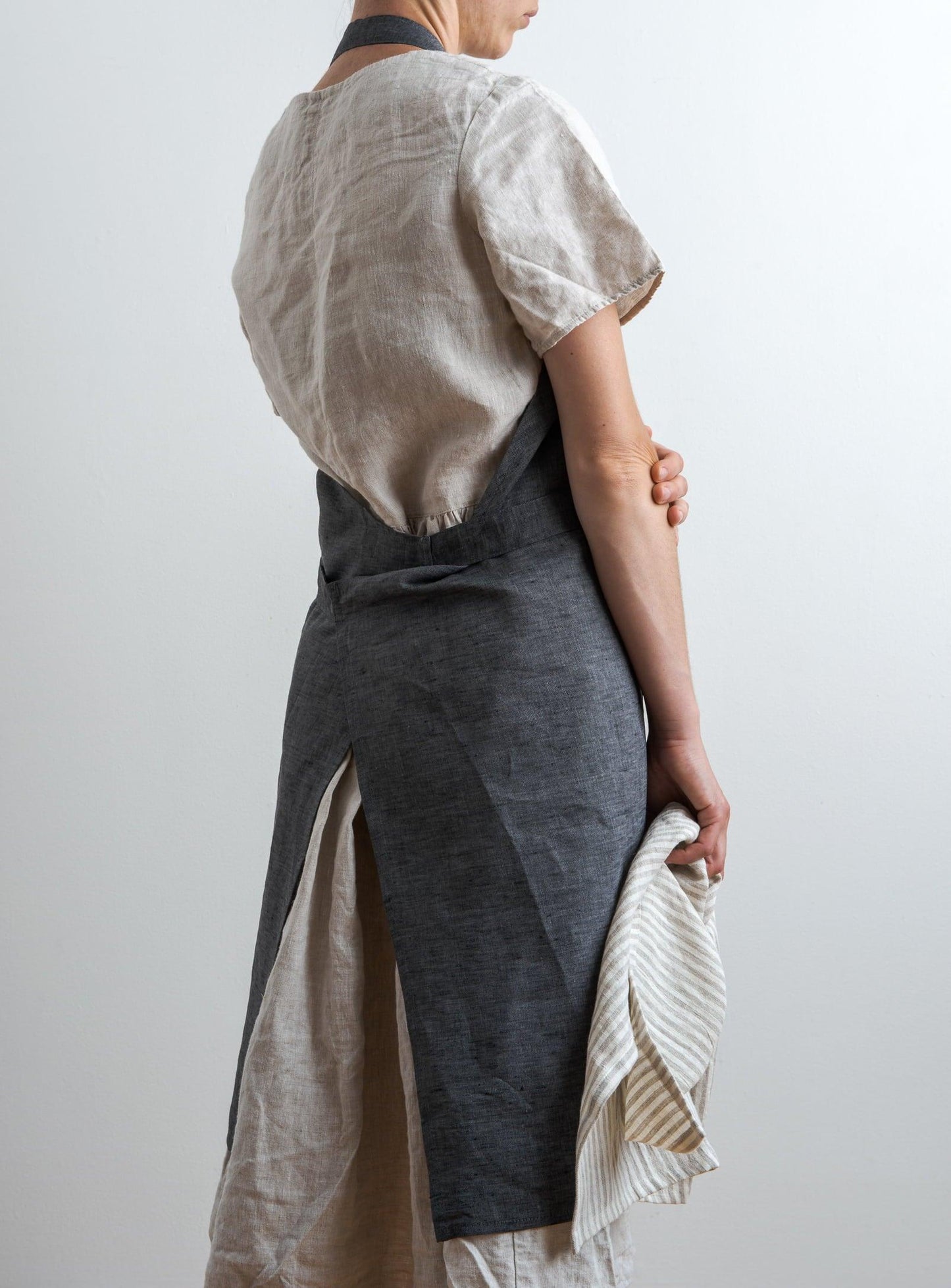 woman seen from behind wearing a grey kitchen apron and holding a striped tea towel